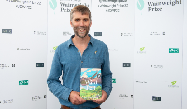 Image of Lee Schofield earing a blue shirt and dark trousers, holding up a copy of his book Wild Fell, in front of a white background with the Wainwright Prize logo on it.